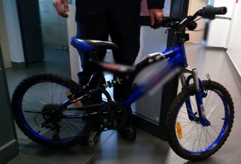 zdjęcie ilustracyjne, obraz przedstawia policjanta skadrowanego od pasa w dół trzymającego rower dziecięcy koloru czarno - niebieskiego