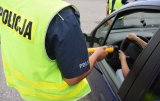 Zdjęcie pokazuje jak policjant w kamizelce odblaskowej bada poziom stężenia alkoholu u osoby siedzącej za kierownicą auta osobowego