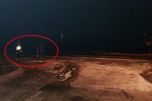 Zdjęcie zrobione nocą, widać jedynie zarys drogi i w oddali z niewielkim odblaskiem postać. Postać zaznaczona jest czerwoną pętlą, aby uzmysłowić odbiorcom jak ważny jest choćby najmniejszy element odblaskowy
