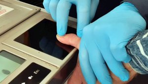 zdjęcie wykadrowane tak, że widać kawałek skanera do daktyloskopii i ręce w niebieskich rękawiczkach pobierające odciski palców