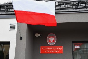Flaga Polski powiewa na wietrze i widać tablicę informacyjną na której widnieje napis Posterunek Policji w Nowogrodzie