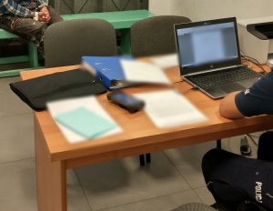 Zdjęcie wykadrowane tak, że widać bok policjanta siedzącego przy biurku... na drugim planie na ławie pod ścianą siedzi osoba z kajdankami na rękach