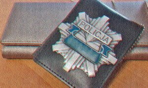 Prostokątny portfel damski koloru jasnego , leżący na stole. na nim oparta jest zawieszka z odznaką policyjną