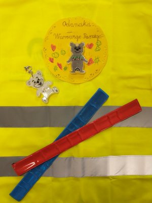 Odznaka wzorowego pieszego wykonana przez dziecko oraz opaski odblaskowe na tle żółtej kamizelki odblaskowej
