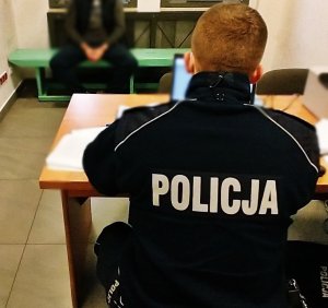 Policjant siedzący przy biurku tyłem do obiektywu, pod ścianą na ławce siedzi mężczyzna z rękoma skutymi w kajdanki