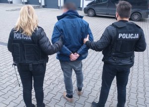 Policjantka i policjant stoją wraz z mężczyzną, który ma założone kajdanki na ręce trzymane z tyłu.