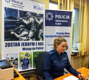 Umundurowana policjantka siedząca przy biurku, za nią plakaty promujące służbę w Policji i Krajową Mapę Zagrożeń Bezpieczeństwa.