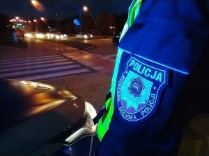 Ulica miasta po zmroku, w tle widać oznakowane przejście dla pieszych, rękaw munduru policyjnego z emblematem Komendy Miejskiej Policji w Łomży.