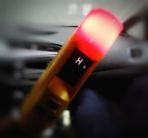 Urządzenie do badania stanu trzeźwości, które nazywa się alcoblow, z czerwoną lampką informującą, iż badana osoba jest pod wpływem alkoholu. W tle wnętrze radiowozu.
