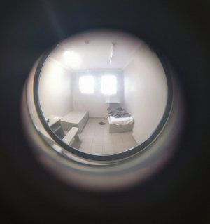 Wizjer w drzwiach do celi, w oddali widać, leżącego mężczyznę oraz dwa zakratowane okna.