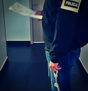 Stojący w korytarzu nieumundurowany policjant. Ma on na ramieniu opaskę z napisem POLICJA w jednej ręce trzyma dokumenty w drugiej kajdanki.