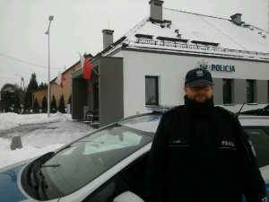 Umundurowany policjant stojący przy radiowozie, w tle budynek Posterunku Policji w Nowogrodzie.
