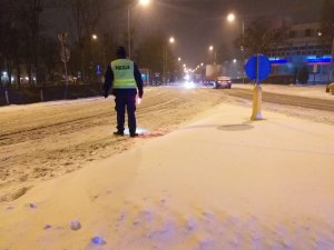 Policjant stojący na jezdni, w ręku trzyma latarkę policyjną z włączonym światłem czerwonym. Kieruję ruchem, jezdnia cała w śniegu. Zdjęcie wykonano porą nocną.
