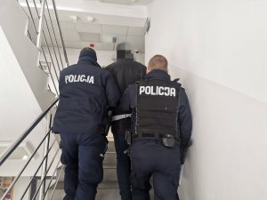 Dwóch policjantów prowadzi zatrzymanego mężczyznę. Zdjęcie wykonano na schodach w budynku komendy.