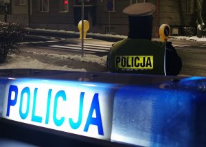 Podświetlony napis na dachu radiowozu Policja oraz stojący tyłem policjant. Panuje wieczór, w oddali ulica miasta Łomży.