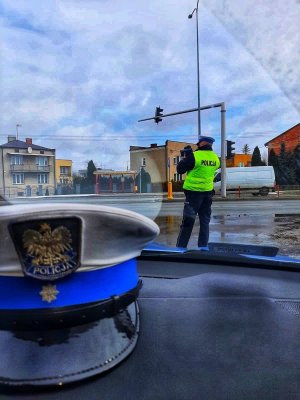 Policjant Ruchu drogowego stojący przy ulicy i mierzący prędkość jazdy uczestników ruchu. Na pierwszym planie policyjna czapka w radiowozie.