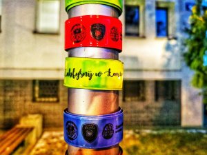 Cztery odblaski w różnych kolorach, zawieszone na rurce od znaku, z napisem Zabłyśnij w Łomży oraz logo Komendy Miejskiej Policji w Łomży, Straży Miejskiej i Urzędu Miasta Łomża.
