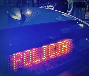 Zdjęcie wykadrowane tak, ze widać napis POLICJA na tylnej szybie w radiowozie i plecy stojącego obok pojazdu funkcjonariusza