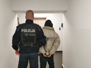 Umundurowany policjant stoi na korytarzu komendy z osobą zatrzymaną. Mężczyzna założone ma kajdanki na ręce trzymane z tyłu.