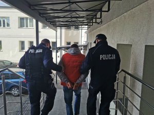 Dwóch umundurowanych policjantów prowadzi jedną osobę zatrzymaną. Mężczyzna ma założone kajdanki na ręce trzymane z tyłu. Zdjęcie wykonano na schodach dziedzińca komendy w Łomży.