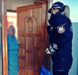 Policjantka stojąca przed drzwiami. W drzwiach stoi starsza kobieta. Policjantka wręcza jej upominek wielkanocny.