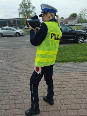 Umundurowana policjantka w kamizelce odblaskowej z napisem policja, stoi przy ulicy i mierzy prędkość jadącym pojazdom ręcznym miernikiem prędkości.
