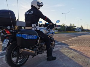 Policjant siedzący na motocyklu - na poboczu drogi. W tle dwupasmowa ulica.