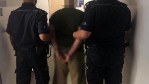 Dwóch umundurowanych policjantów trzyma osobę zatrzymaną, która ma założone kajdanki na ręce trzymane z tyłu. Stoją w korytarzu komendy.