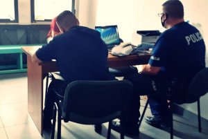 Policjanci w pomieszczeniu, siedzą przy biurku na którym są dokumenty, pod oknem na drewnianej ławie siedzi mężczyzna