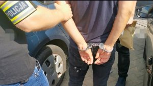 Ręce zatrzymanego w kajdankach założonych z tyłu. Widoczna ręka policjant, na której założona jest opaska z napisem policja.