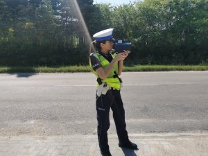 Umundurowana policjantka stoi przy drodze i mierzy prędkość ręcznym miernikiem prędkości.
