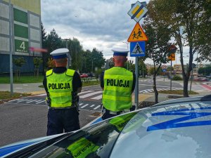 Dwóch umundurowanych policjantów w kamizelkach odblaskowych stoją przy oznakowanym przejściu dla pieszych.