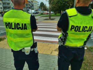 Dwóch umundurowanych policjantów w kamizelkach odblaskowych stoją przy oznakowanym przejściu dla pieszych.
