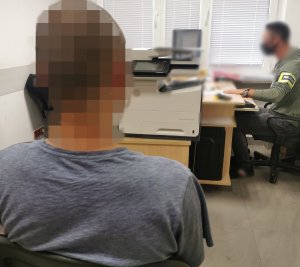 Policjant w ubraniu cywilnym z założoną opaską z napisem Policja siedzi przy komputerze i wykonuje czynności służbowe z zatrzymanym, który siedzi na krześle.