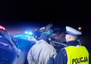 Umundurowany policjant trzyma osobę zatrzymaną. W tle ciągnik rolniczy przewrócony w rowie oraz radiowóz policyjny z włączonymi światłami błyskowymi.