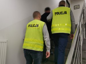 Policjanci w ubraniach cywilnych w kamizelkach odblaskowych z napisem Policja prowadzą po schodach zatrzymanego mężczyznę.
