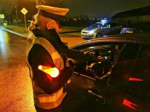 Umundurowana policjantla w kamizelce odblaskowej z napisem policja przeprowadza kontrolę stanu trzeźwości kierowcy.