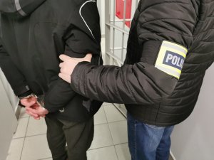 Policjant w ubraniu cywilnym, z opaska z napisem policja trzyma zatrzymanego, który ma założone kajdanki na ręce trzymane z tyłu.