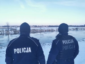 Dwóch policjantów stojących tyłem, a tle rzeka, po której pływają kawałki lodu.