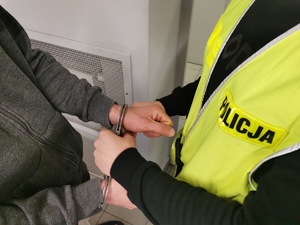 Policjant w ubraniu cywilnym w kamizelce z napisem policja zakłada kajdanki zatrzymanemu mężczyźnie