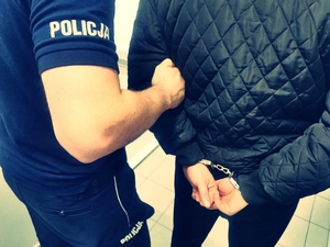 Policjant prowadzi zatrzymanego, który trzyma ręce z tyłu i ma na nich kajdanki