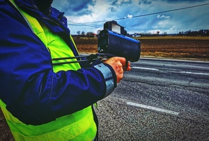 Policjant trzymający ręczny miernik prędkości. Na zdjęciu widać też drogę i pola