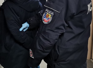 Mundur policyjny z emblematem Komendy Miejskiej Policji w Łomży, dłoń policjanta w rękawiczce jednorazowej. Policjant trzyma osobę zatrzymaną.