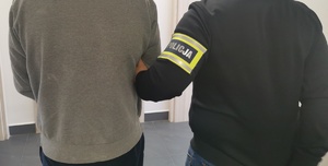 Policjant w ubraniu cywilnym z opaską na ręku z napisem policja trzyma zatrzymanego mężczyznę, który ma założone kajdanki.