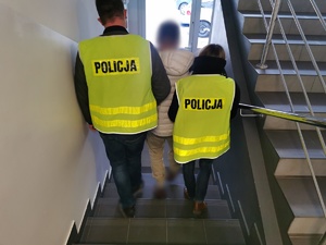 Policjanci w ubraniach cywilnych z kamizelkami z napisem policja prowadzą schodami zatrzymanego mężczyznę.