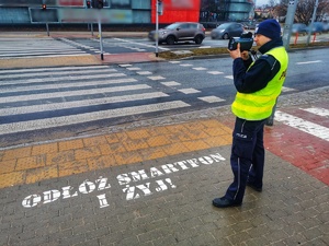 Umundurowany policjant stoi przy jezdni i mierzy prędkość kierowcom.