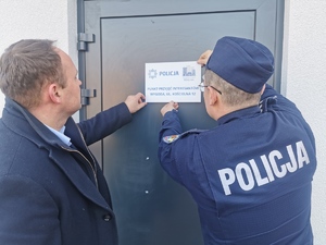 Komendant Miejski Policji w Łomży wspólnie z Wójtem Gminy Łomża przyklejają szyld na drzwiach, informujący o punkcie przyjęć interesantów.