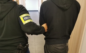Policjant w ubraniu cywilnym z opaską na ręku z napisem policja trzyma zatrzymanego mężczyznę.