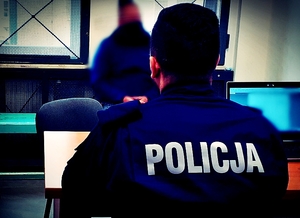 Policjant siedzący przy biurku. Po przeciwnej stronie pod ścianą z oknem siedzi mężczyzna.