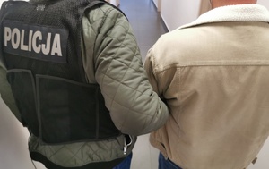 Policjant w ubraniu cywilnym w kamizelce z napisem policja trzyma zatrzymanego mężczyznę.